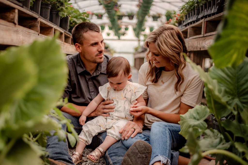 A family portrait of a mom, dad, and toddler sitting surrounded by foliage in a greenhouse.