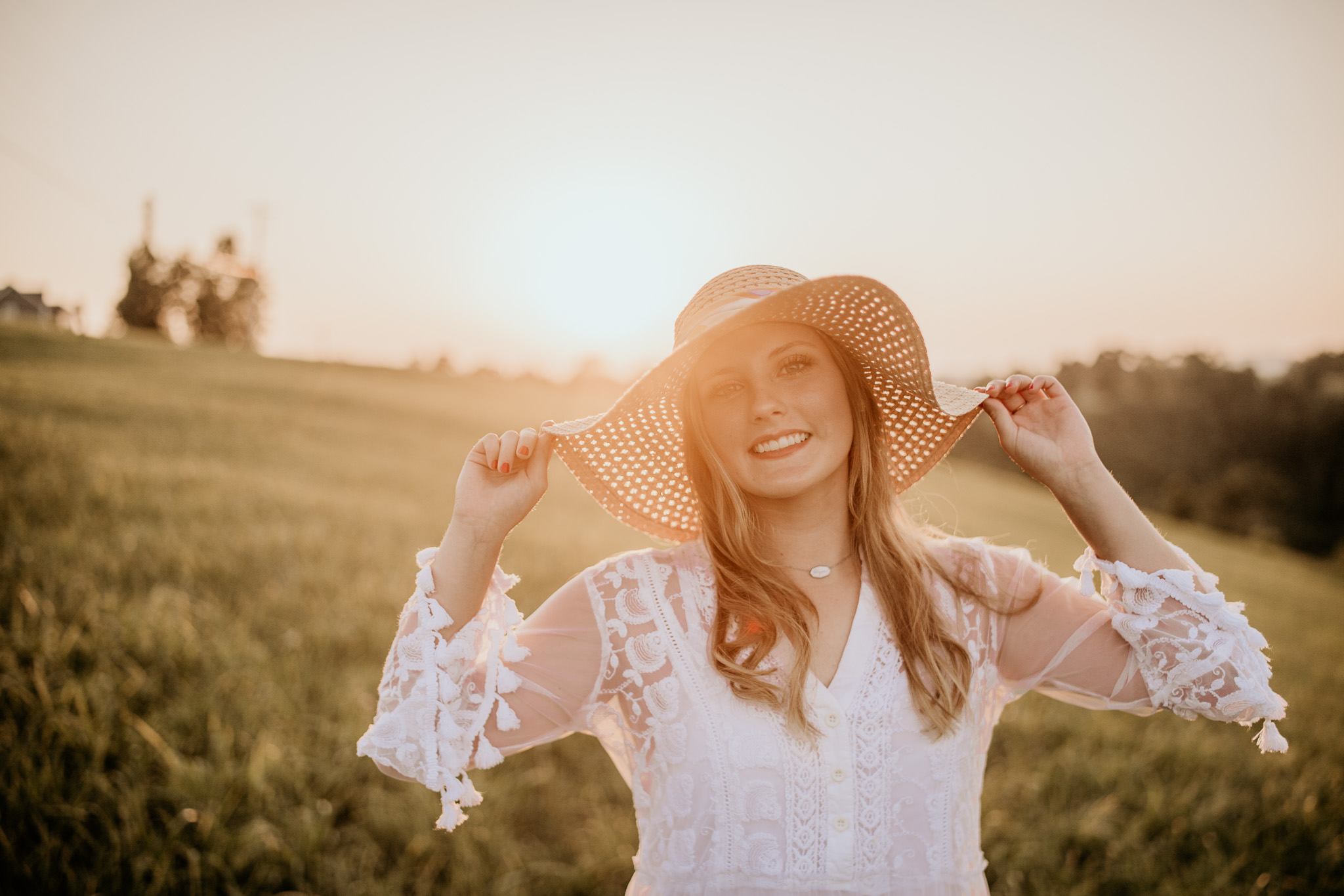 A senior portrait of a young woman standing in  the last rays of the sun's golden hour holding the edges of her hat.